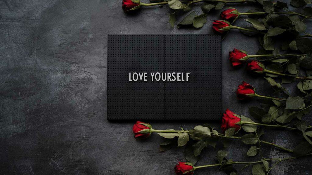 A black mat with the words love yourself written on it. Red roses surround the mat. Symbolizes self-care in recovery.
