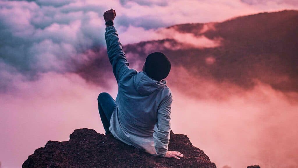 Young man with sitting on a mountain top with his left arm raised above his head in a fist symbolizing strength in addiction recovery.